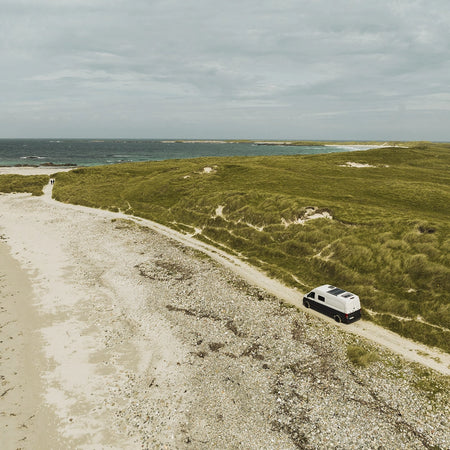 Chewie, Roamer's campervan travelling along a road on the beachfront in Scotland. Beautiful white sands and turquoise waters.