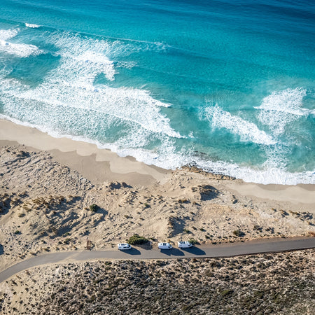 3 sprinter campervans in convoy driving along a road at the edge of a beach. Beautiful turquoise seas and white sandy beaches.