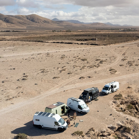 5 sprinter campervans parked up in a remote, sandy location with mountain views. A group of people socialising around a table outside of the vans.